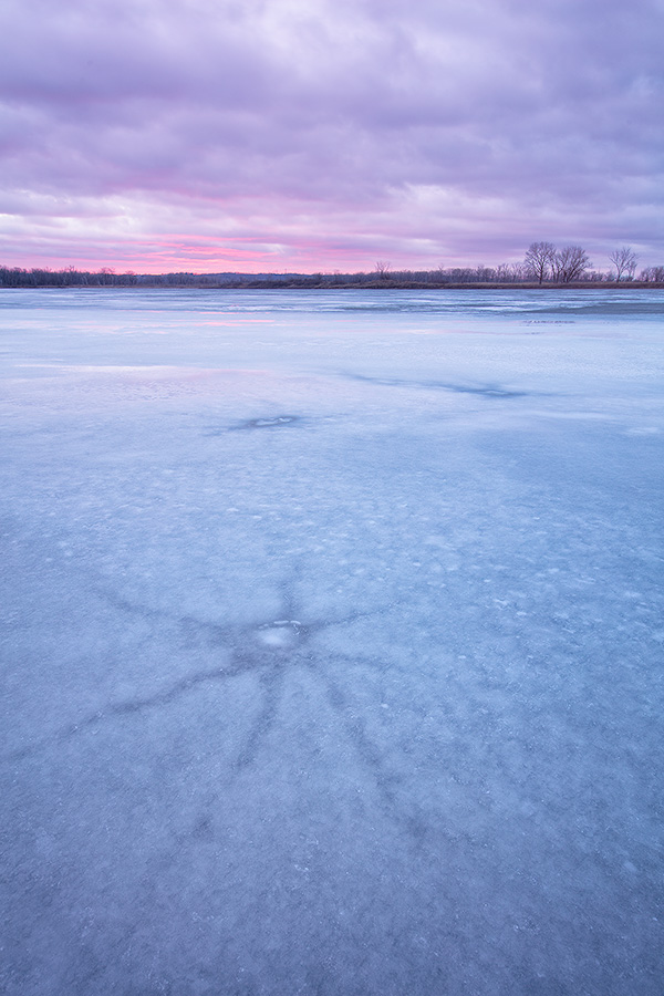 On this evening at DeSoto National Wildlife Refuge a quiet stillness prevailed.  I wandered the shore of the lake looking for interesting patterns in the now melting ice.  I found some interesting star shapes that had emerged from the recent thaws.  Sunset was near and although it was cloudy, I was hoping for a little illumination of the clouds.  After a while I gave up and begin leaving when a bit of sun broke free and I quickly set up again and captured this image. - DeSoto Photography
