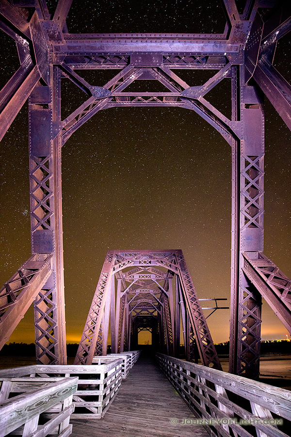 On a clear night at Niobrara State Park, I setup on the railroad trussle converted to a footbridge near the confluence of the Niobrara and Missouri Rivers.  Using a flashlight I lightpainted the bridge while exposing for the stars.  In the distance the lights of Niobrara can be seen glowing on the horizon. - Nebraska Photography