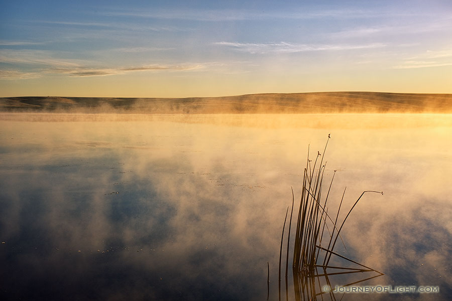One of the most expansive and uninhabited areas of Nebraska also holds expansive beauty.  The Oglala National Grasslands in northwestern Nebraska represent some of the last native grasslands in the United States.  On a cool September morning a few years ago, I ventured out to the Meng Reservoir and captured the morning sun hitting the mist rising from the water.  The only sound was ducks quacking the distance welcoming the new day. - Nebraska Photography